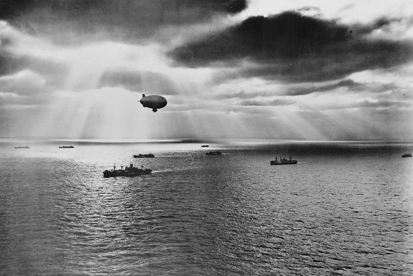 A U.S. Navy blimp hovers over a peaceful convoy moving across the Atlantic during World War II, scanning the waters for enemy submarines, June 1943.