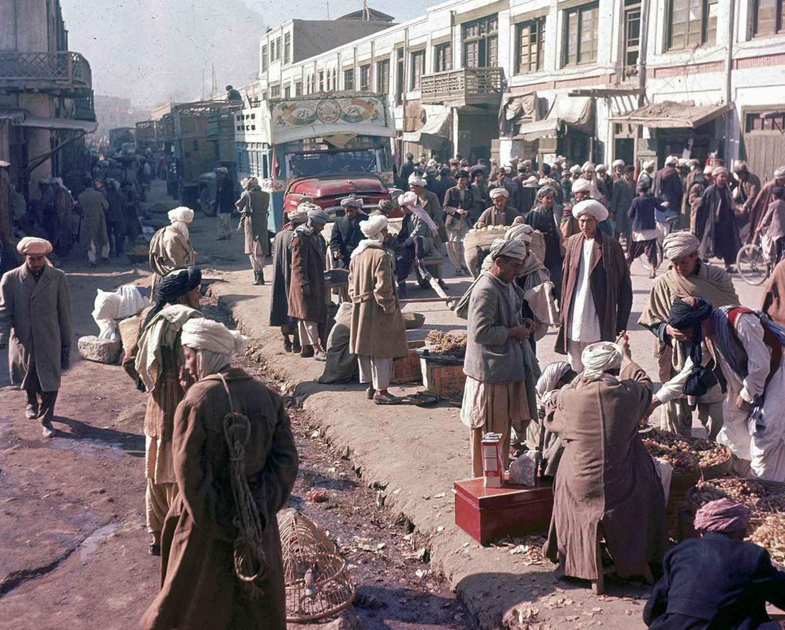 A bustling Kabul street in November 1961, where men stroll past roadside vendors as a vibrantly painted truck passes by.