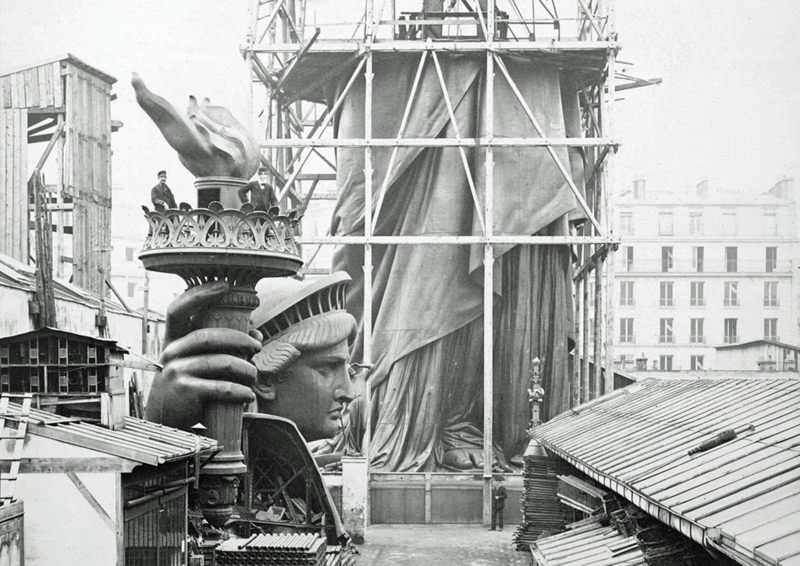 The lower half of the Statue of Liberty stands under scaffolding in Paris, with its detached head and torch at its feet, photographed in 1883.