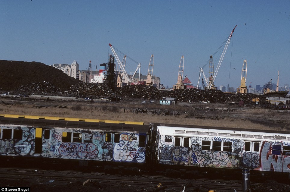 A New York subway train, fully covered in graffiti, halts amidst a backdrop of construction cranes and urban decay.