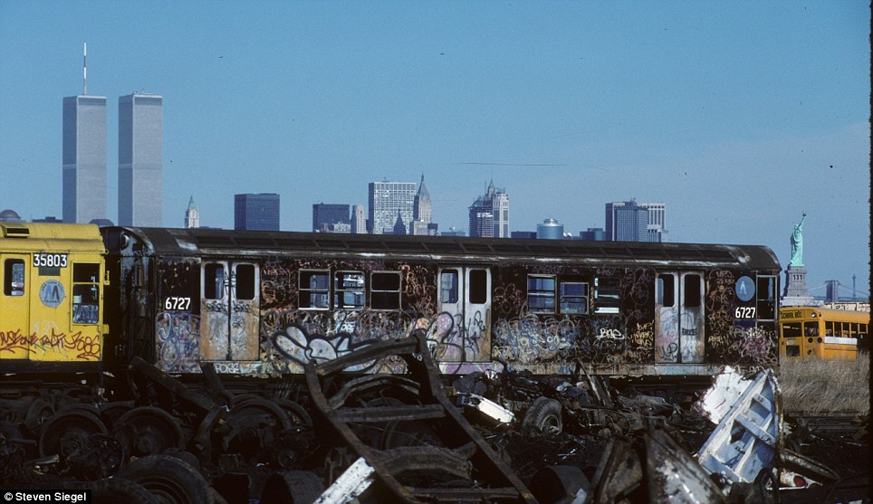 A vivid contrast: a scrapyard in the foreground, while the iconic New York skyline, with the Statue of Liberty, shines brightly in the background.